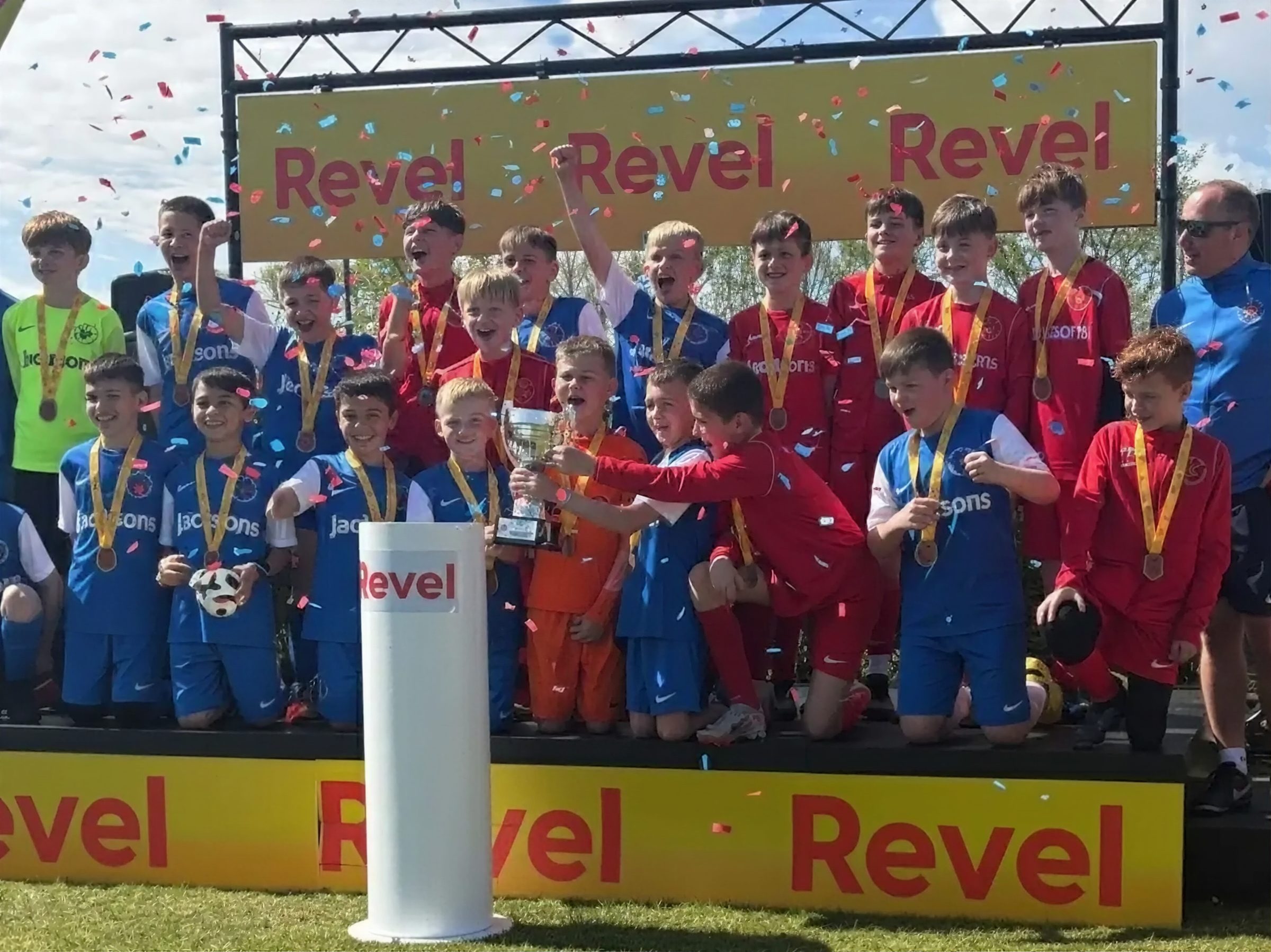 A squad lifting the cup on the Revel podium under falling confetti, medals round every neck